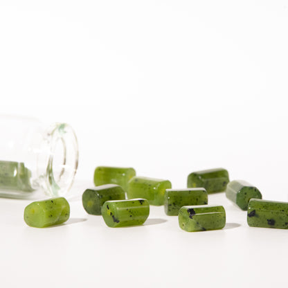 Green rectangular gemstone beads with dark flecks, shown spilling from a clear glass jar onto a white surface against a bright white background.