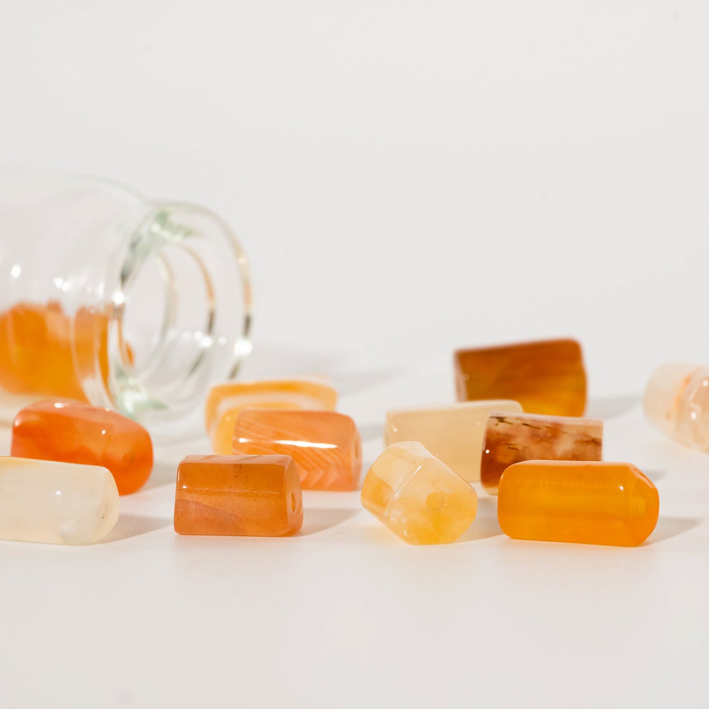 Several small polished stone beads in orange, amber, cream, and translucent tones scattered on a white surface, with a tipped-over clear glass bottle in the blurred background.