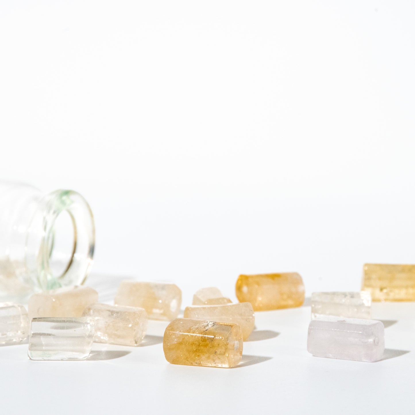 Several translucent rectangular gemstone-like pieces in pale yellow, clear, and light pink are scattered on a white surface beside a tipped-over clear glass bottle.