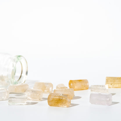 Several translucent rectangular gemstone-like pieces in pale yellow, clear, and light pink are scattered on a white surface beside a tipped-over clear glass bottle.