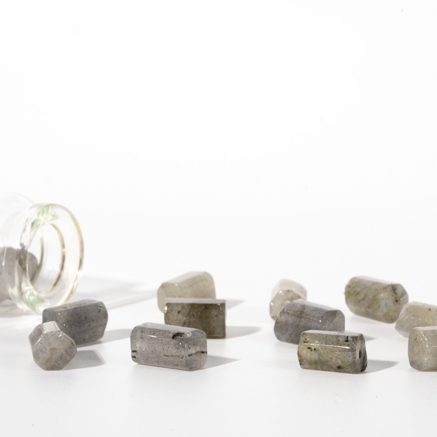 Several small gray polished stone pucks are scattered on a white surface, with a tipped-over clear glass container on the left, photographed in bright studio light.