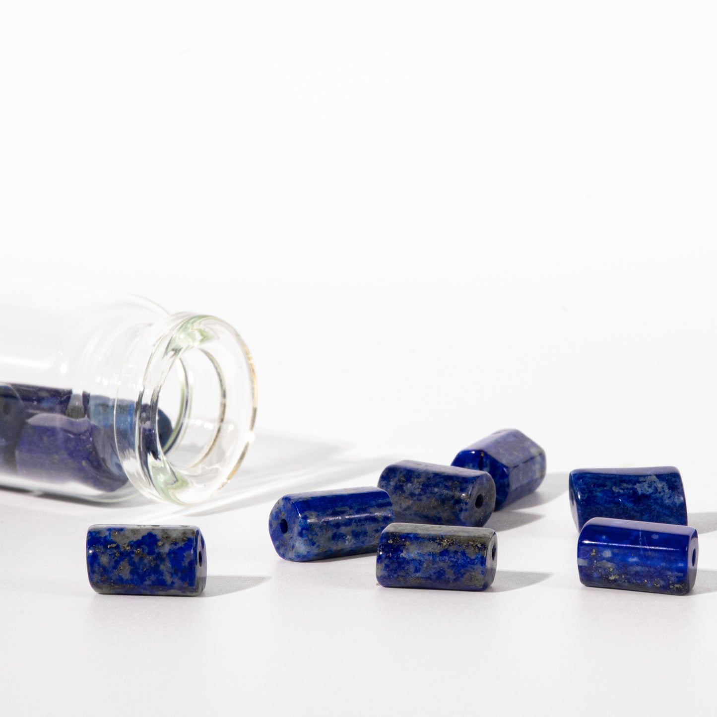 Several polished lapis lazuli cube beads with natural golden inclusions are scattered on a white surface next to a tipped-over clear glass jar filled with more beads.