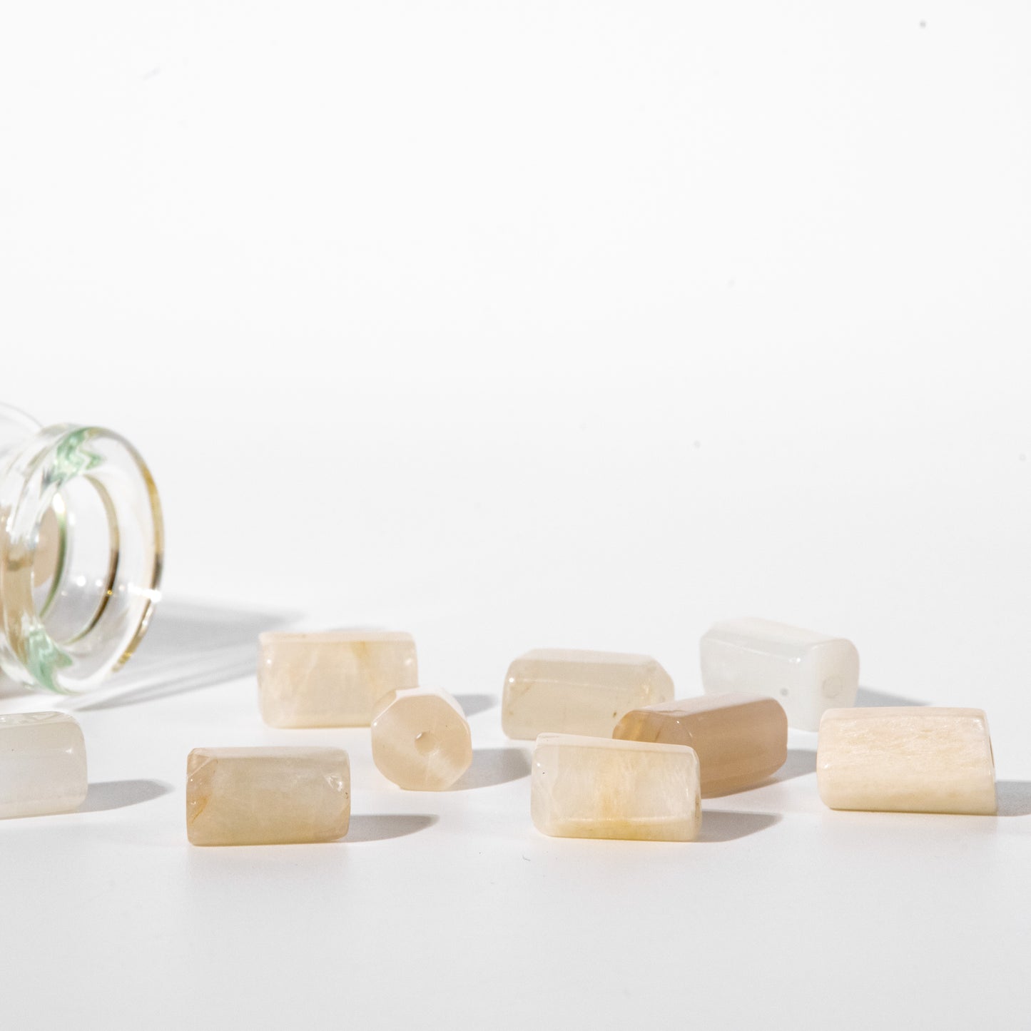 Several small translucent pale cream rectangular beads or stone pieces scattered on a white studio surface, with a cropped clear glass container visible on the left.