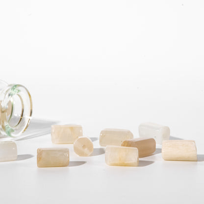 Several small translucent pale cream rectangular beads or stone pieces scattered on a white studio surface, with a cropped clear glass container visible on the left.