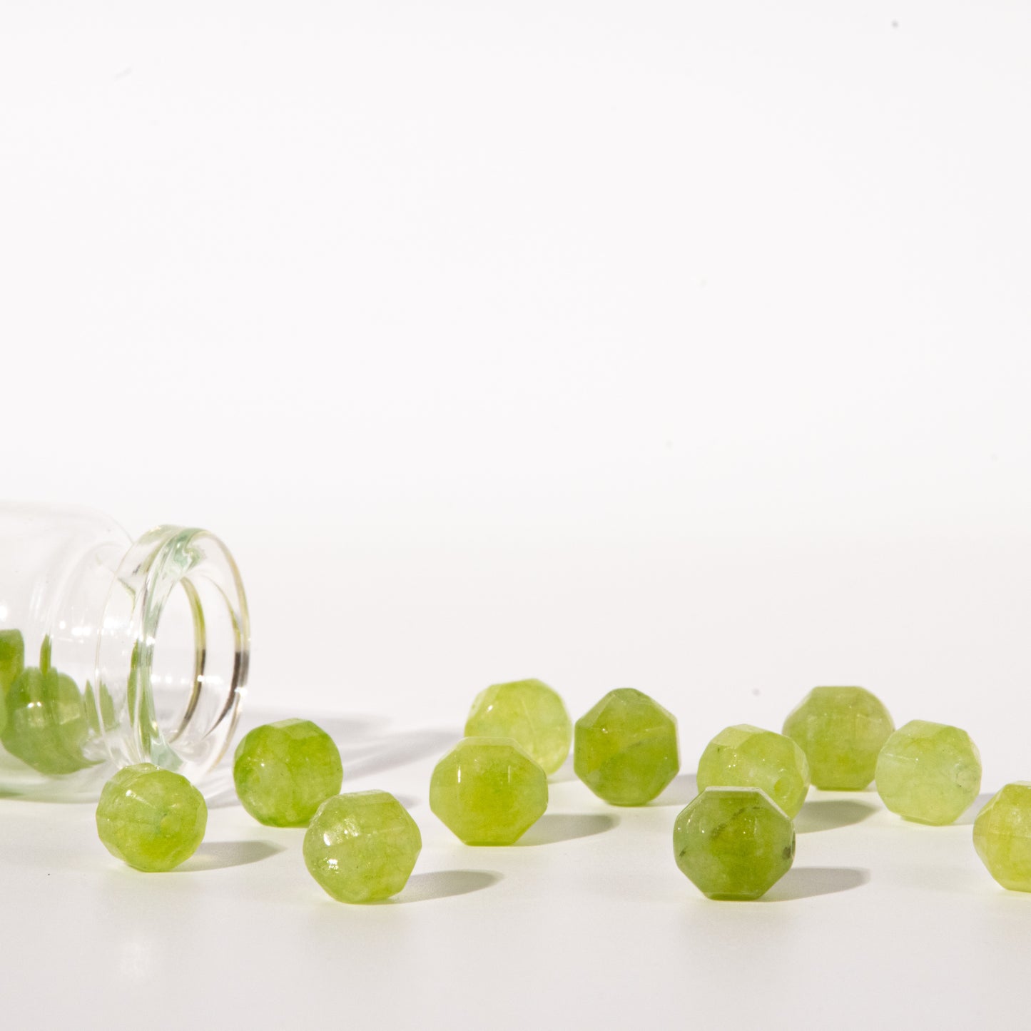 Faceted light green gemstone beads spilling from a clear glass bottle onto a white surface against a white background.