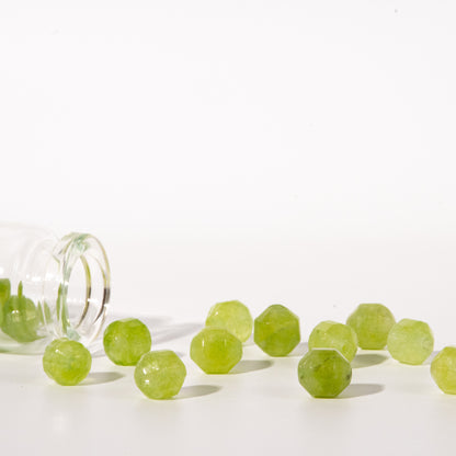 Faceted light green gemstone beads spilling from a clear glass bottle onto a white surface against a white background.