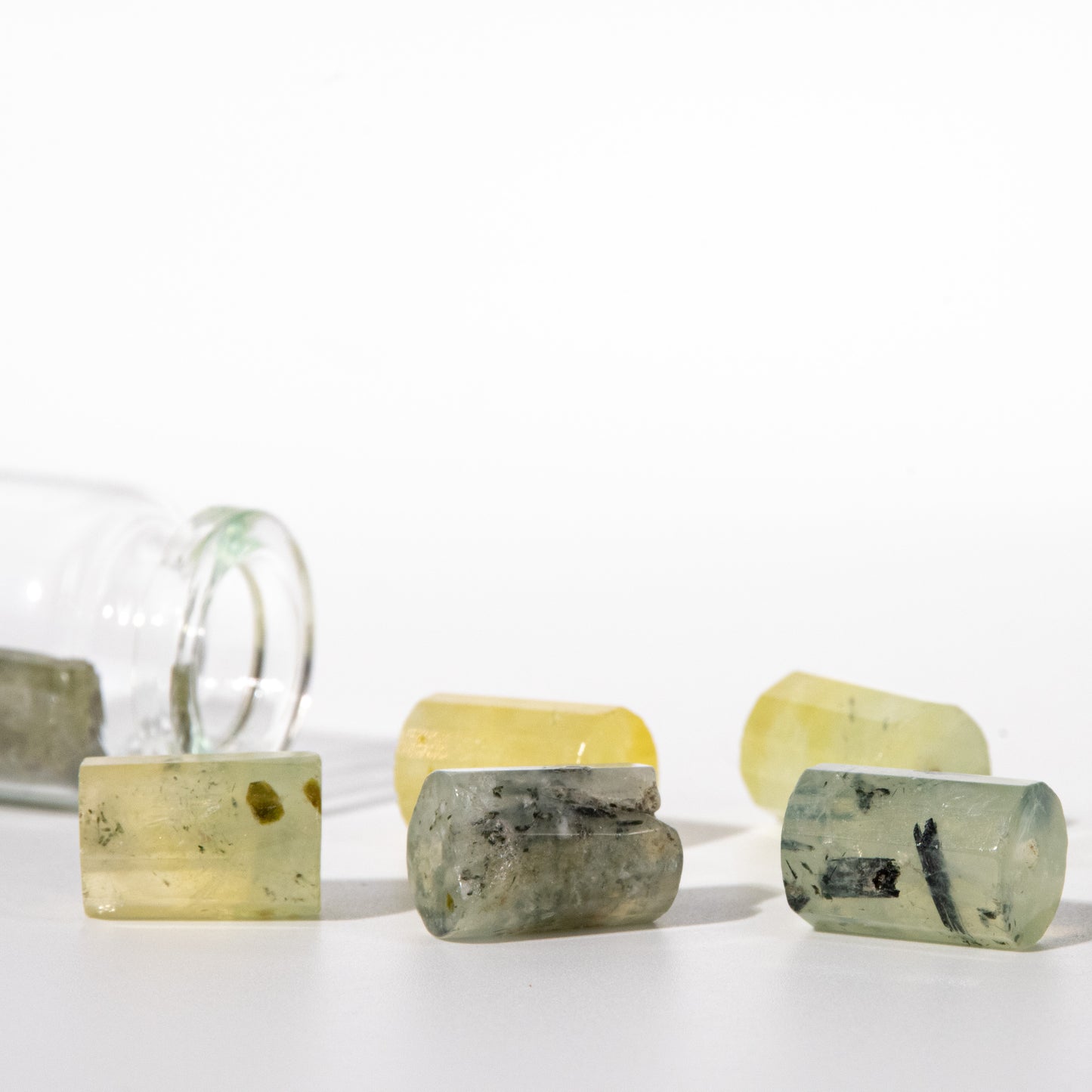 Several small translucent gemstone-like cylinders and rectangular pieces in pale yellow, green, and gray tones are arranged on a white surface, with a clear glass bottle lying on its side in the background.