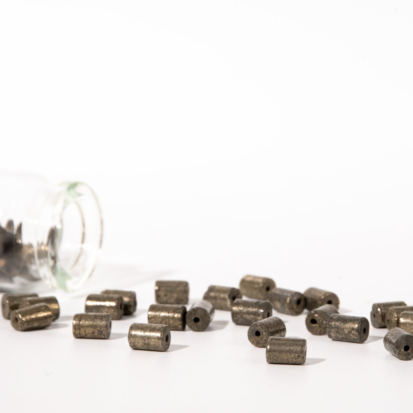 Small metallic cylindrical beads with a matte bronze finish scattered on a white surface, with a clear glass jar tipped over on the left side.
