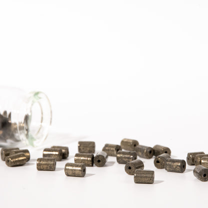 Small metallic cylindrical beads with a matte bronze finish scattered on a white surface, with a clear glass jar tipped over on the left side.