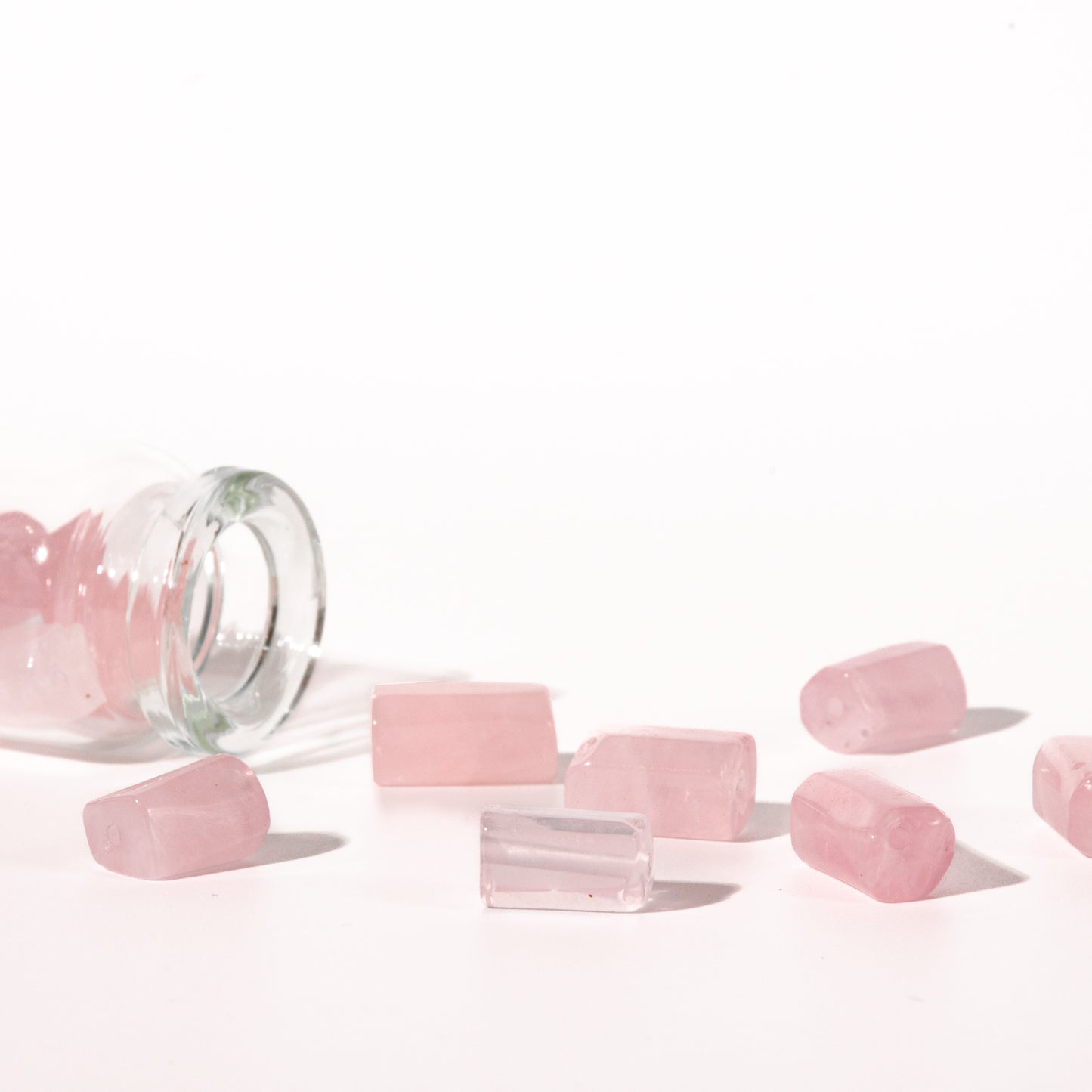 Pink rose quartz crystal chips scattered on a white surface next to a tipped-over clear glass bottle.
