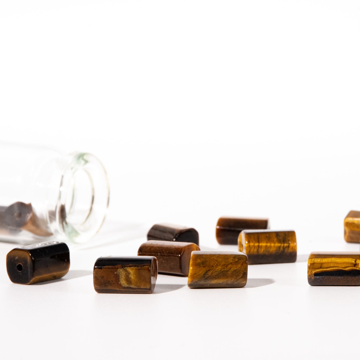 Several rectangular tiger's eye beads with a glossy finish are scattered on a white surface, with one clear glass bottle tipped over in the background on the left.