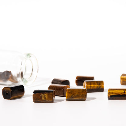 Several rectangular tiger's eye beads with a glossy finish are scattered on a white surface, with one clear glass bottle tipped over in the background on the left.