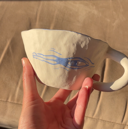 A hand holding a white ceramic cup with a blue abstract eye design, set against a warm, sunlit beige fabric background.