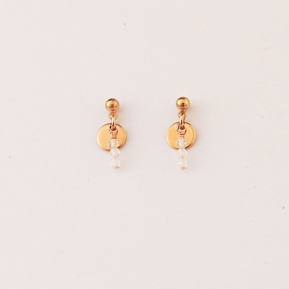 A pair of gold dangle earrings with a small round disc, a tiny gold bead, and a clear faceted bead hanging below, shown on a plain off-white background.