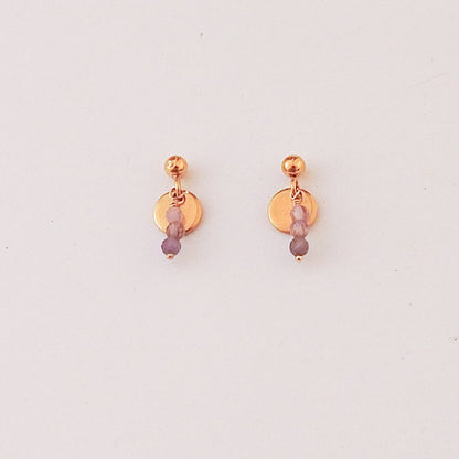 A pair of rose gold stud earrings with a flat circular disc, a small bead, and three dangling faceted beads in lavender and light purple, shown on a plain off-white background.