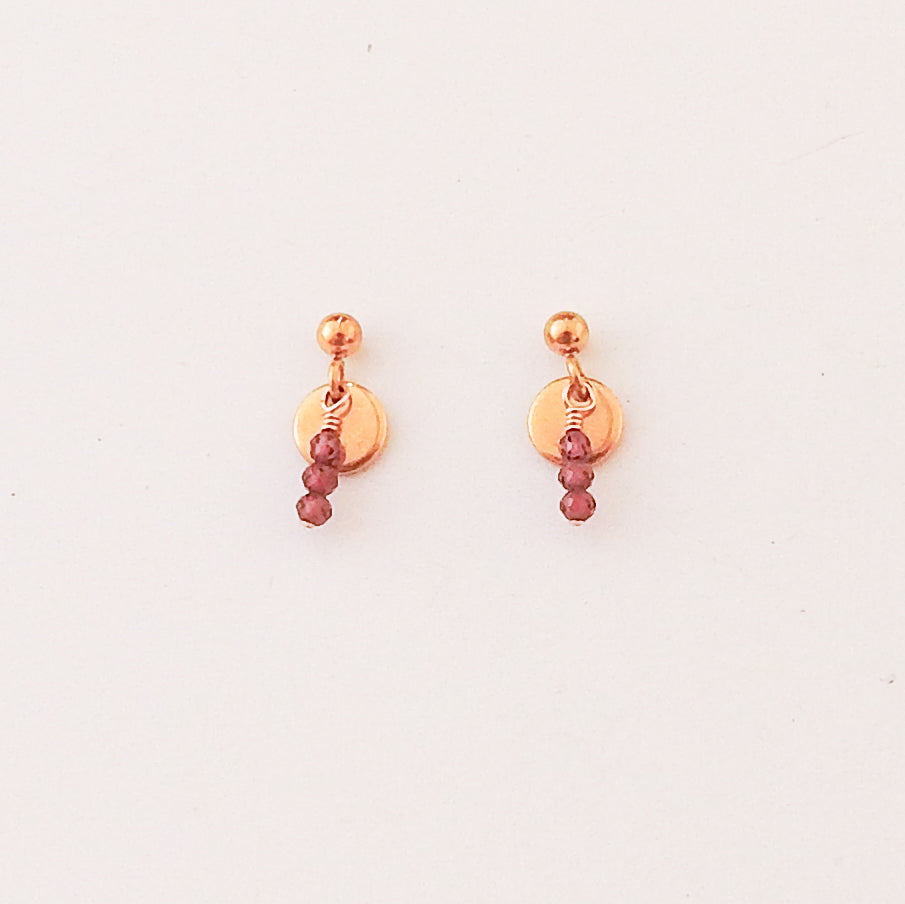 A pair of rose gold dangle earrings with a small round disc, a tiny bead, and three dangling faceted burgundy beads, shown on a plain white background.