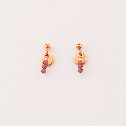 A pair of rose gold dangle earrings with a small round disc, a tiny bead, and three dangling faceted burgundy beads, shown on a plain white background.