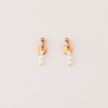 A pair of gold-plated dangle earrings featuring a small round disc, a tiny gold bead, and three small white pearl-like beads hanging down, set against a plain off-white background.