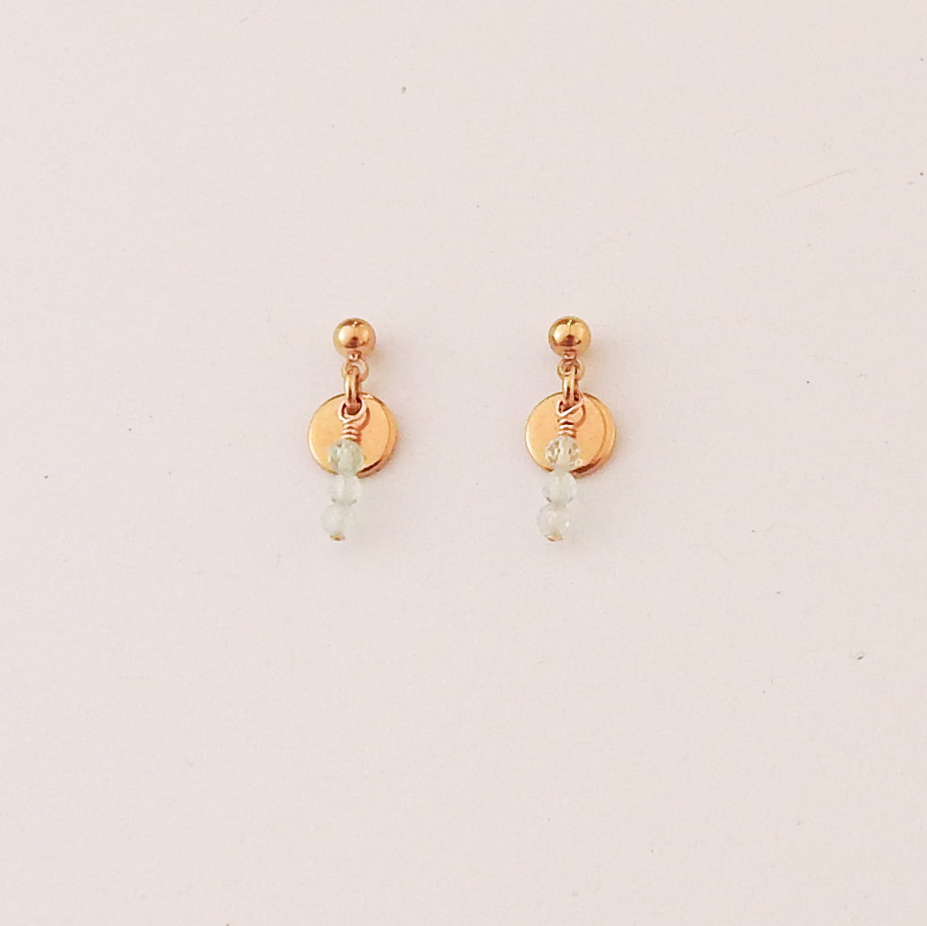 A pair of small rose gold dangle earrings with a flat circular disc, a wrapped bead, and a tiny light blue bead drop, shown on a plain off-white background.