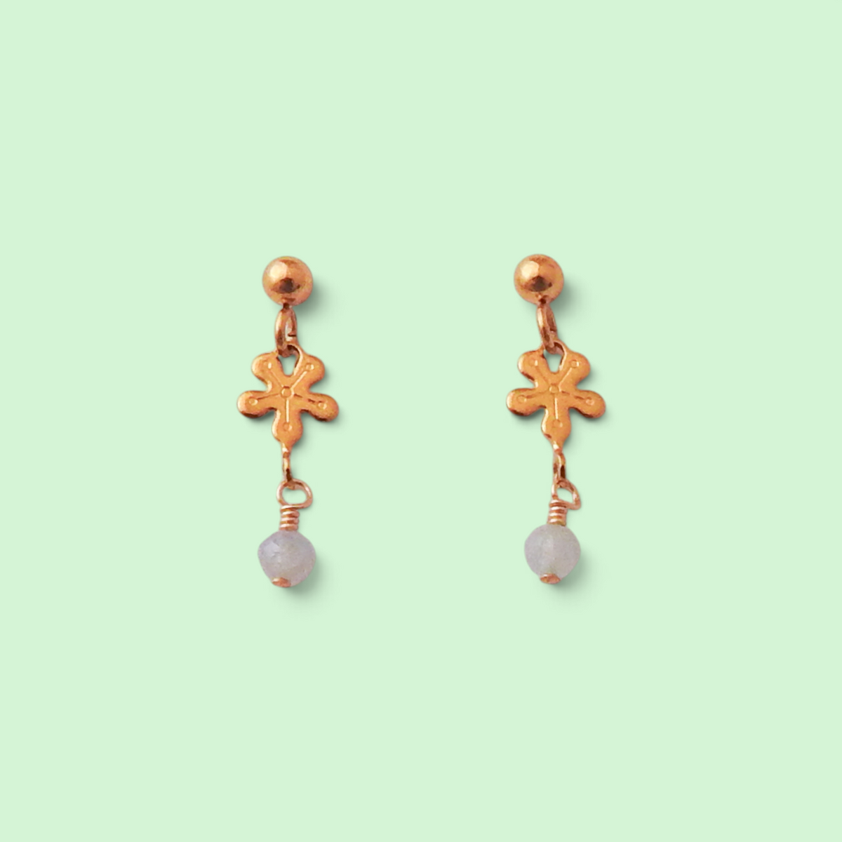A pair of rose gold dangle earrings with a small cross charm, a round stud, and a dangling light purple bead, set against a solid pale green background.