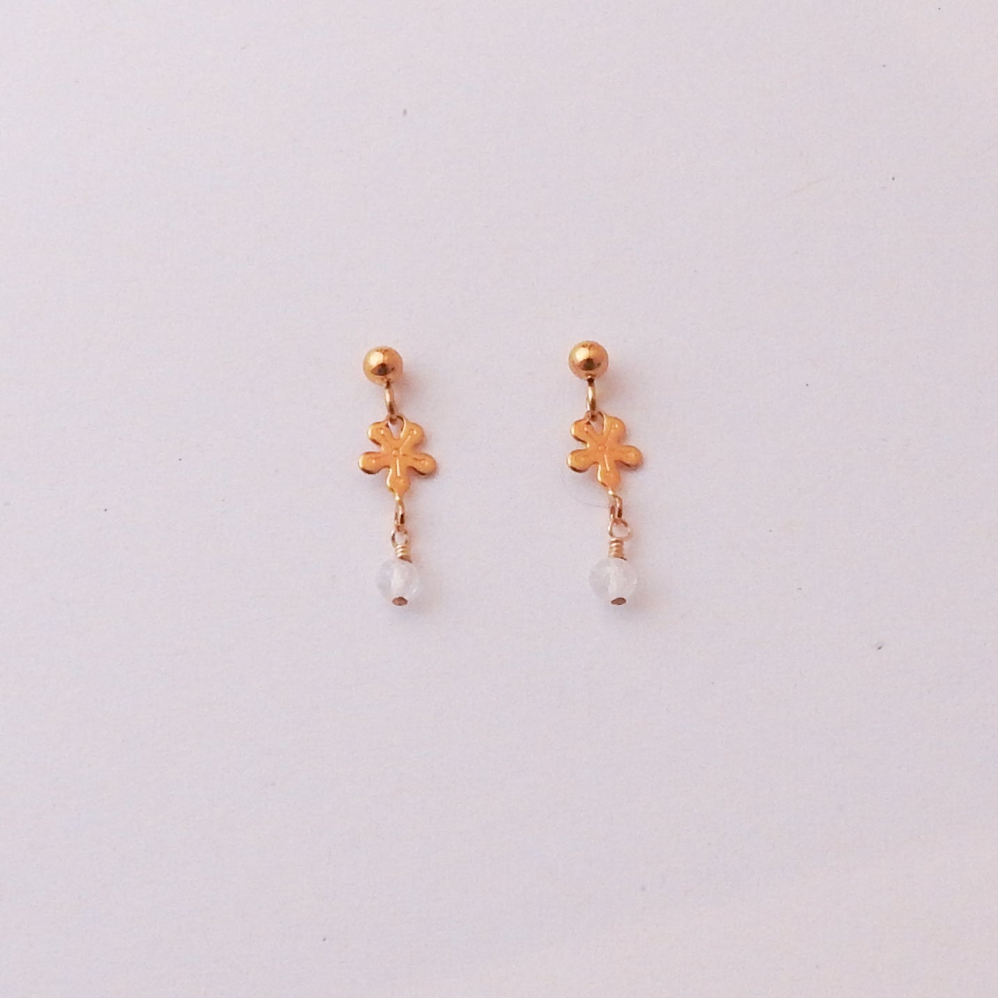 A pair of gold dangle earrings featuring a small flower-shaped charm and a clear faceted bead at the bottom, displayed on a plain light background.