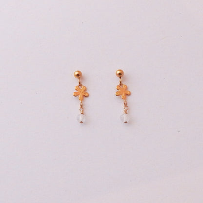 A pair of gold dangle earrings featuring a small flower-shaped charm and a clear faceted bead at the bottom, displayed on a plain light background.