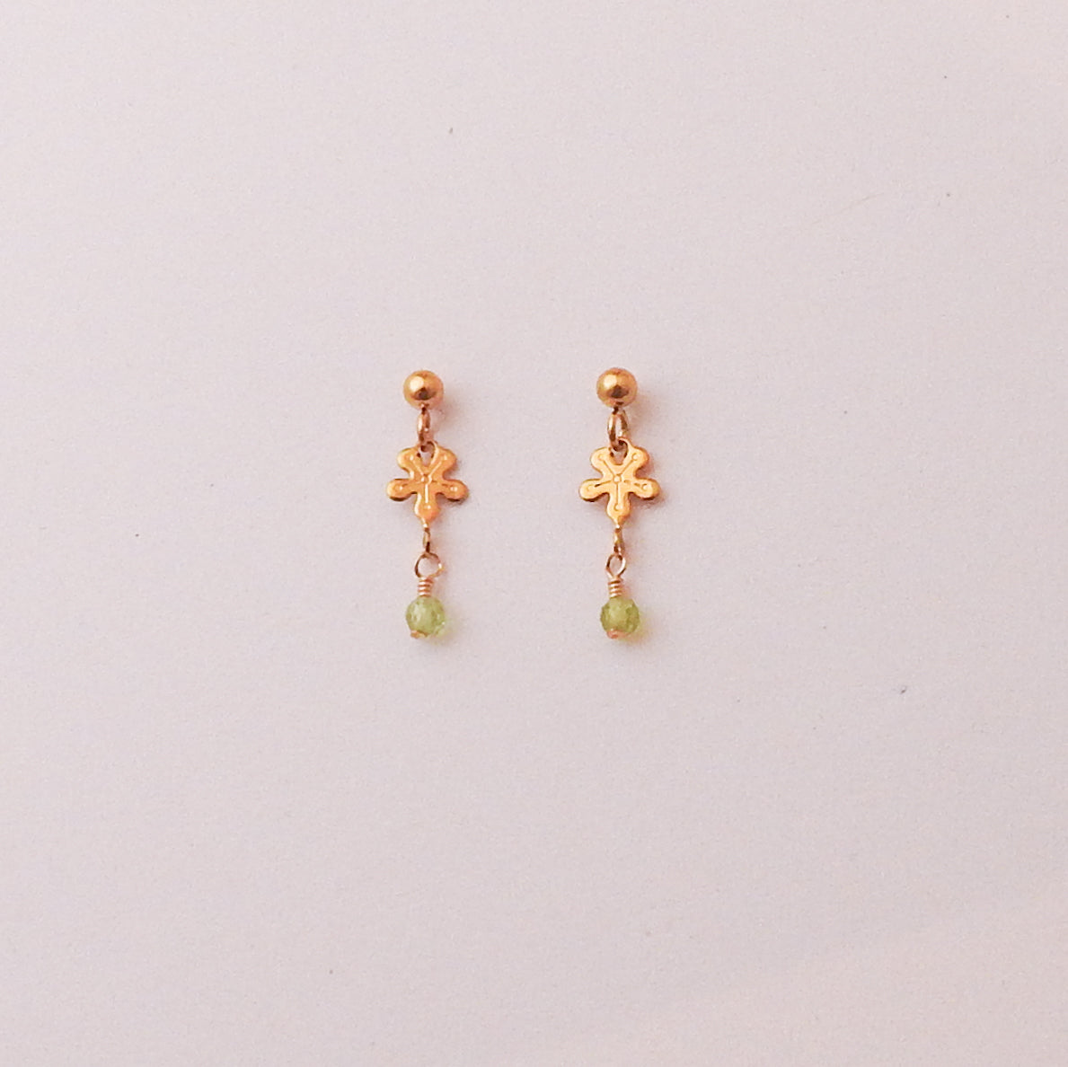 A pair of gold dangle earrings with a small flower-shaped charm, a gold ball stud, and a tiny light green bead dangling below, shown on a plain light background.