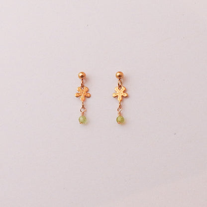 A pair of gold dangle earrings with a small flower-shaped charm, a gold ball stud, and a tiny light green bead dangling below, shown on a plain light background.