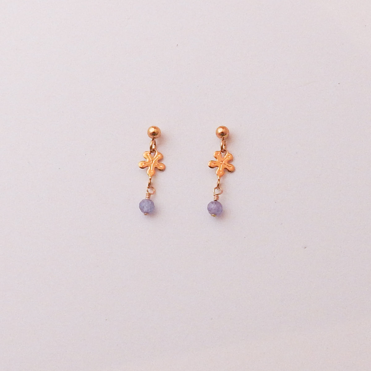 A pair of small gold dangle earrings with a textured flower charm and a small round purple bead dangling below, set against a plain off-white background.