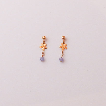 A pair of small gold dangle earrings with a textured flower charm and a small round purple bead dangling below, set against a plain off-white background.