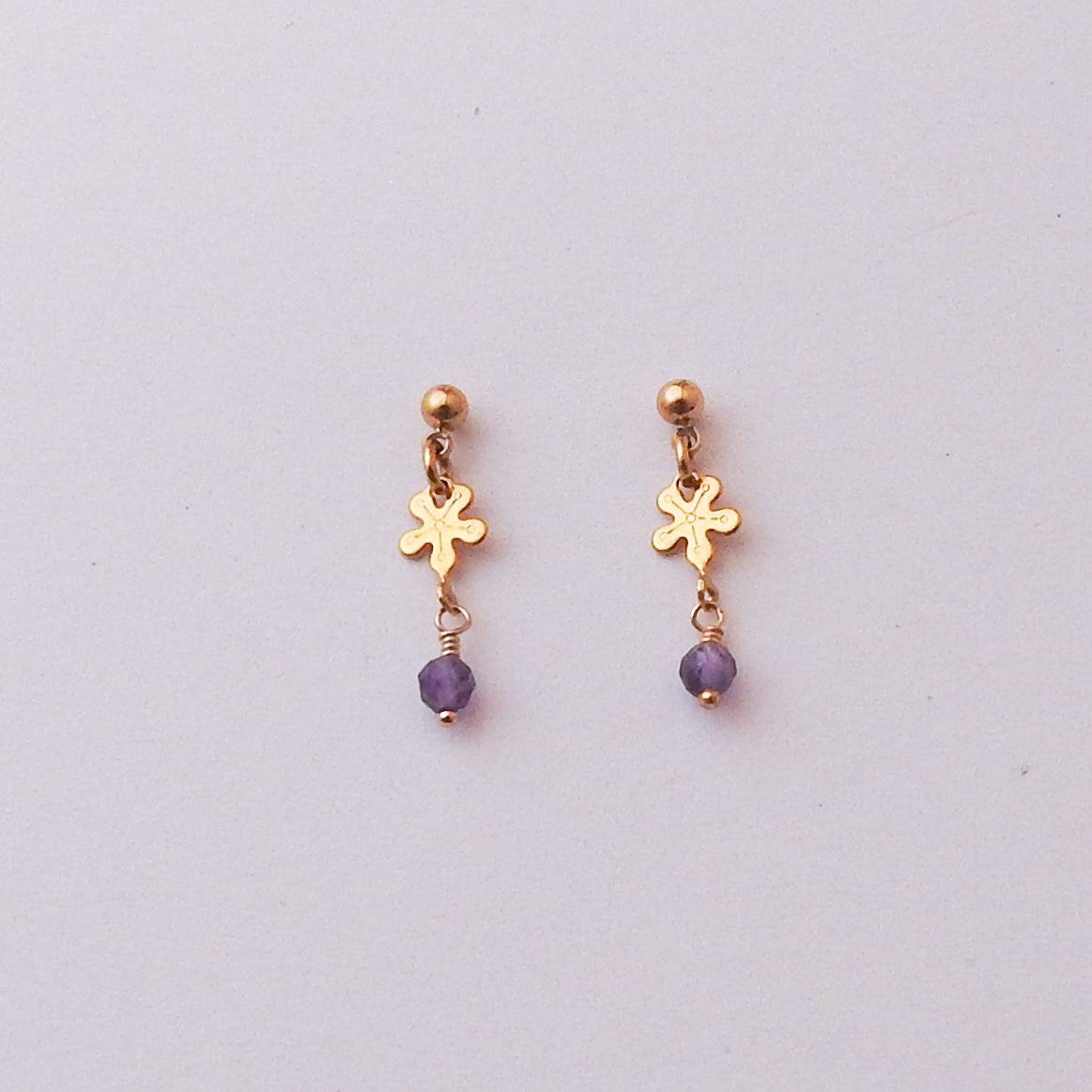 A pair of gold dangle earrings with a small flower-shaped charm, a gold ball stud at the top, and a dangling purple faceted bead, shown on a plain light gray background.