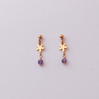A pair of gold dangle earrings with a small flower-shaped charm, a gold ball stud at the top, and a dangling purple faceted bead, shown on a plain light gray background.