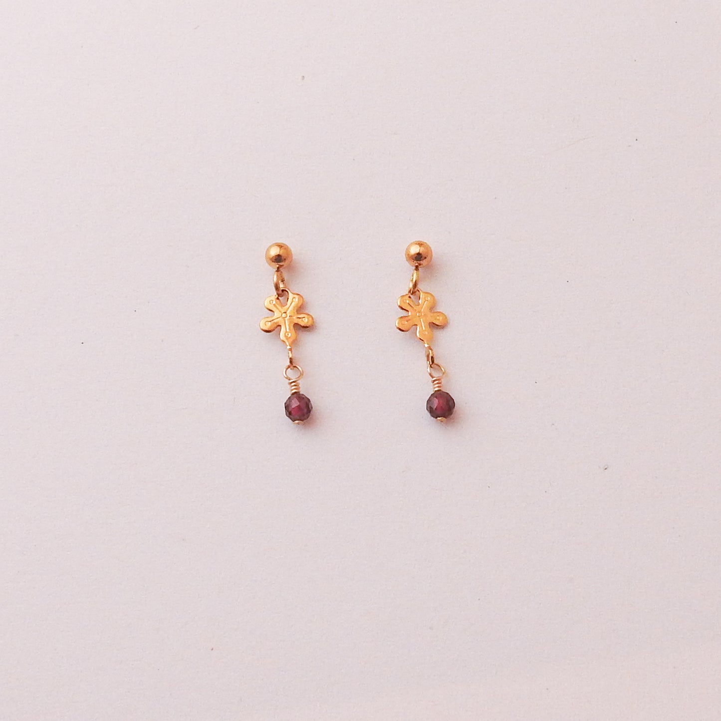 A pair of gold dangle earrings with a small gold charm and a dangling dark red bead, shown on a plain light background.