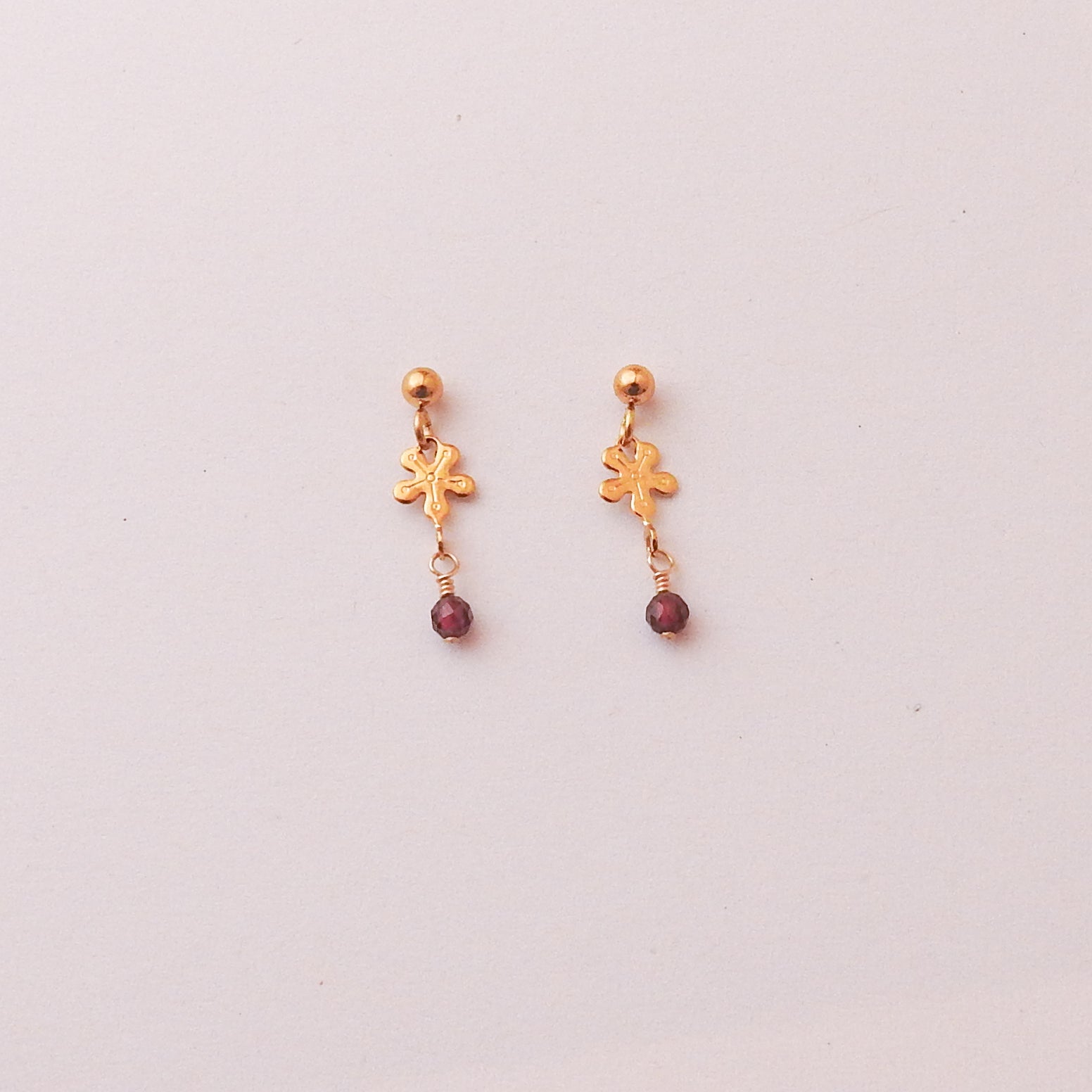 A pair of gold dangle earrings with a small gold charm and a dangling dark red bead, shown on a plain light background.