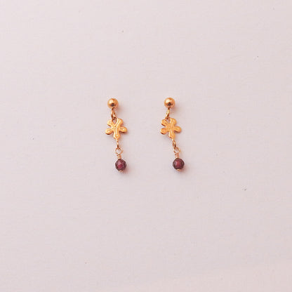 A pair of gold dangle earrings with a small gold charm and a dangling dark red bead, shown on a plain light background.