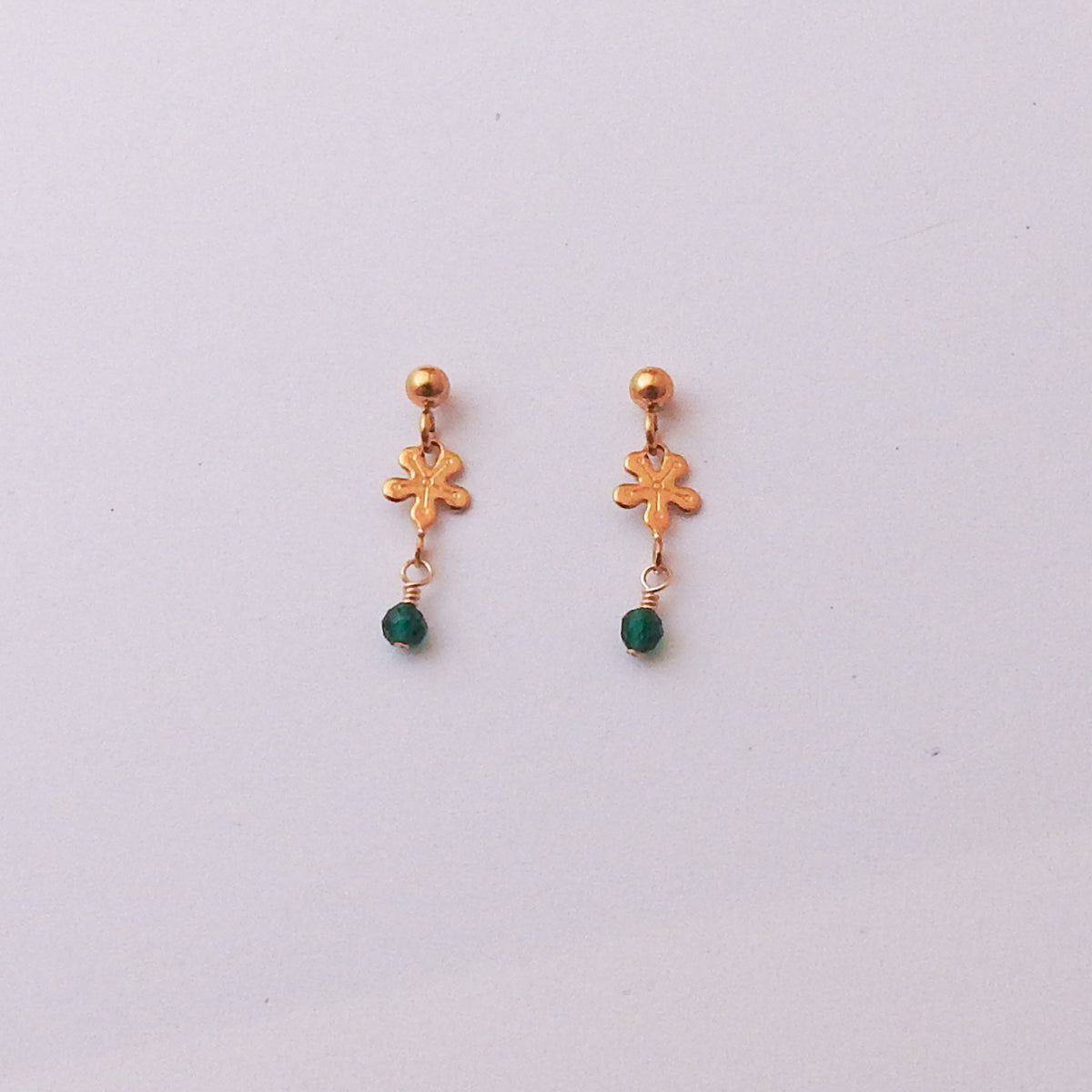 A pair of gold dangle earrings with a small gold floral charm, a tiny gold ball at the top, and a small green bead dangling below, shown on a plain light gray background.