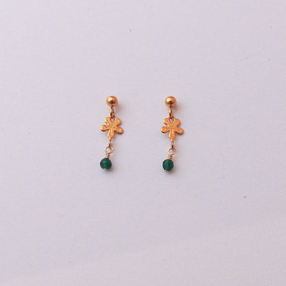 A pair of gold dangle earrings with a small gold floral charm, a tiny gold ball at the top, and a small green bead dangling below, shown on a plain light gray background.