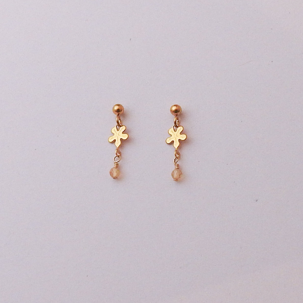 A pair of small gold dangle earrings with a flower-shaped charm, a tiny gold bead above, and a dangling faceted bead, shown on a plain light background.