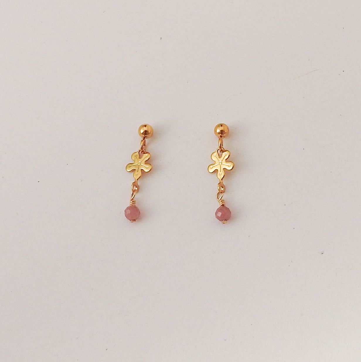 A pair of gold dangle earrings with a small flower charm and a pink bead drop, displayed on a plain light gray background.