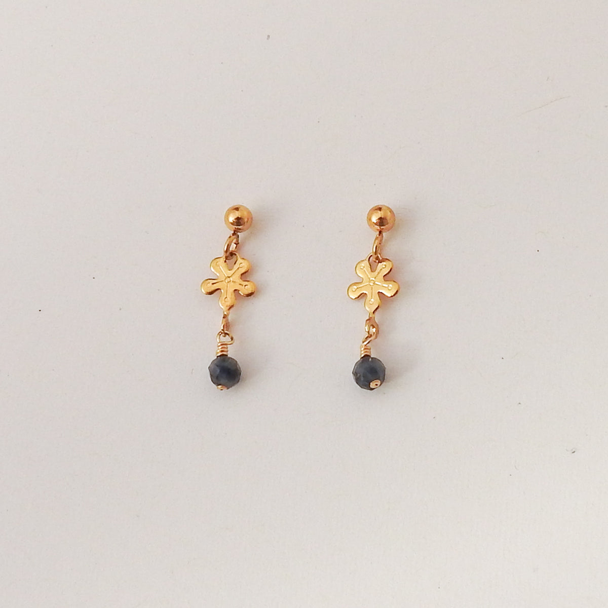 A pair of gold dangle earrings with a small textured cross charm and a dark gray bead drop, shown on a plain light gray background.