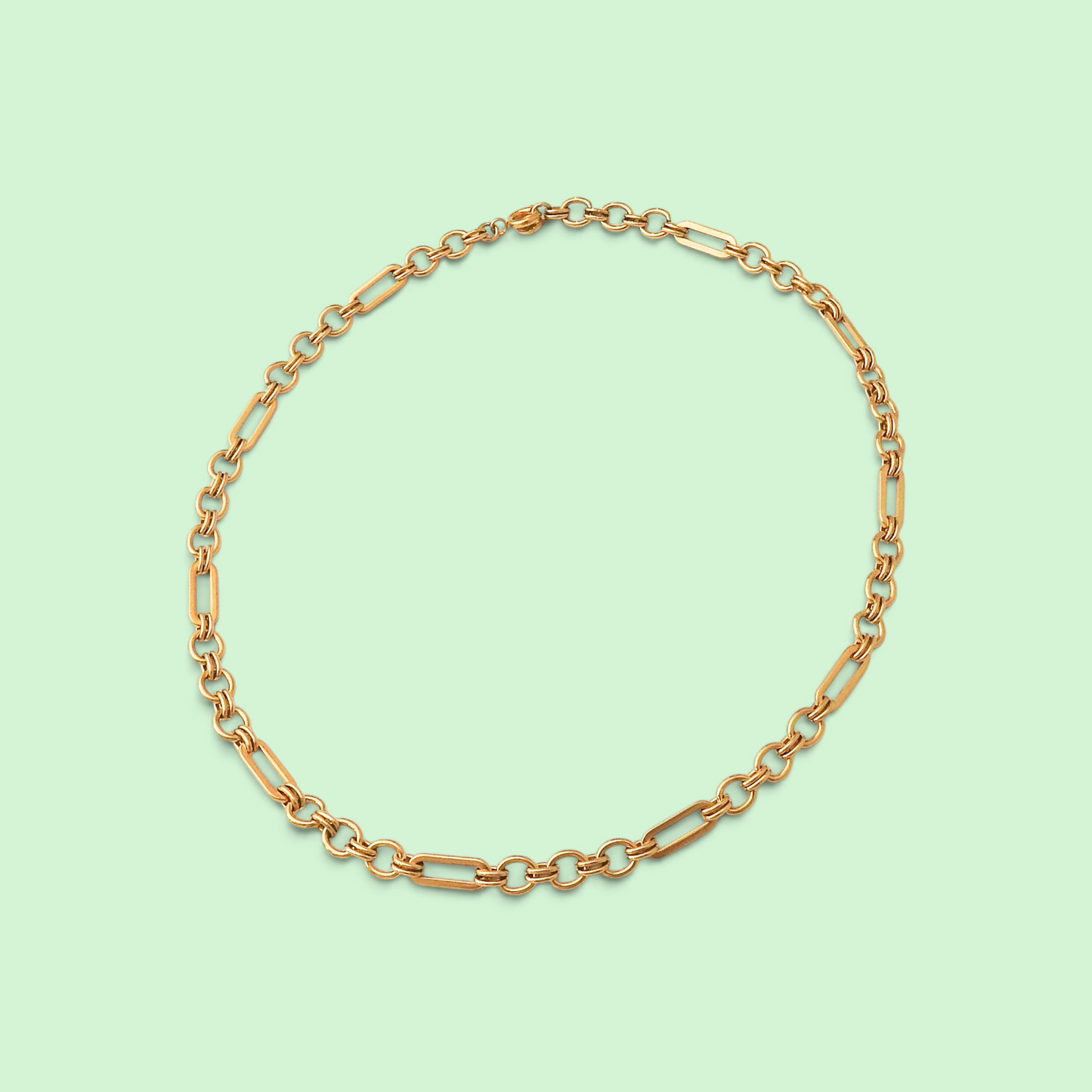 A gold chain necklace with alternating elongated oval links and smaller round links, arranged in a circle on a solid pale mint green background.