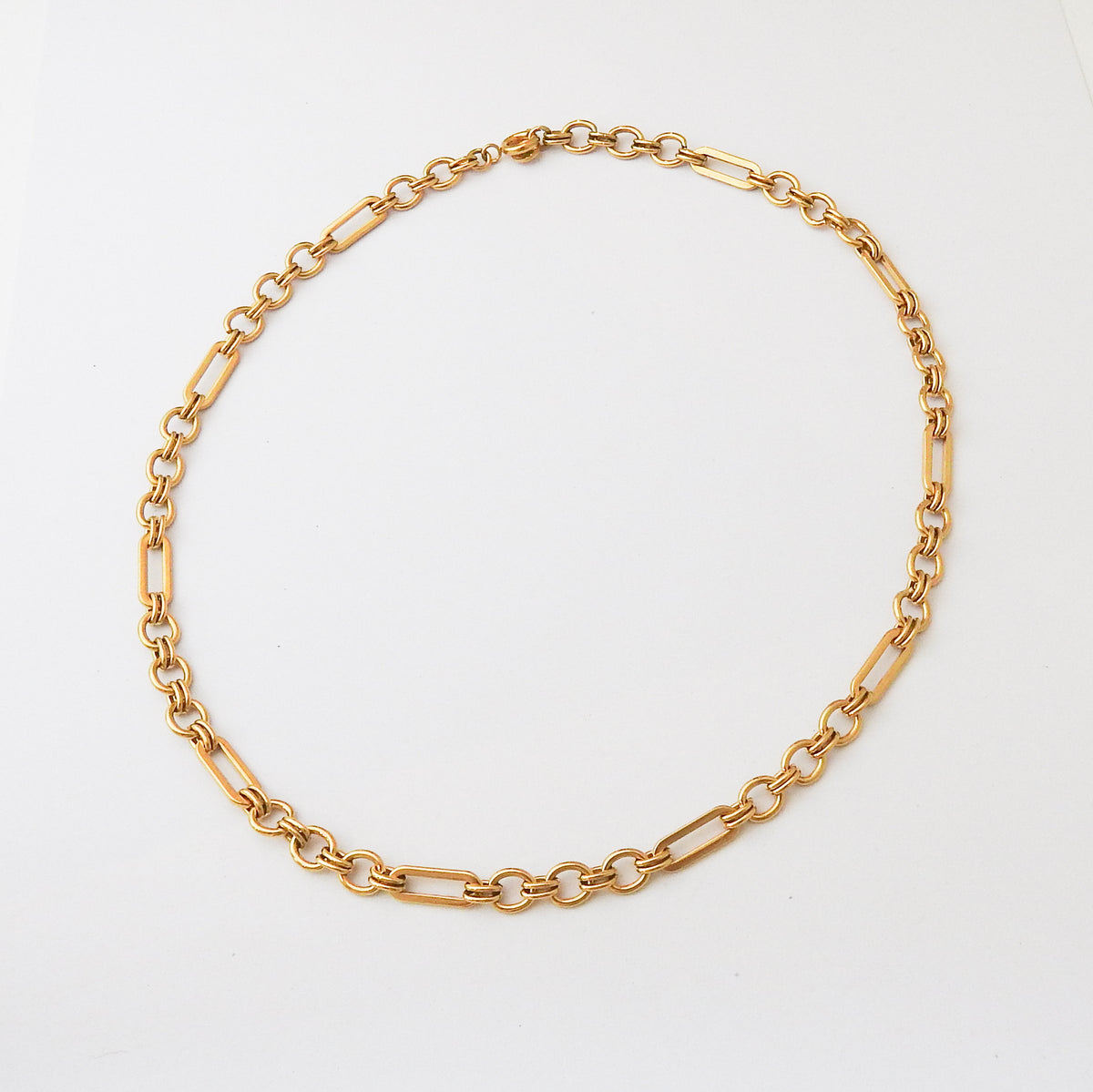 A gold chain necklace with a mix of elongated oval links and smaller circular links, arranged in a circle on a plain white background.