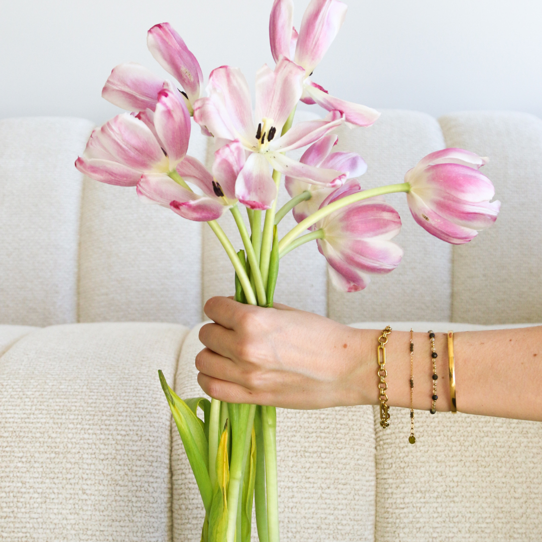 A hand holding a bouquet of pink tulips in front of a light beige textured sofa, wearing gold bracelets.