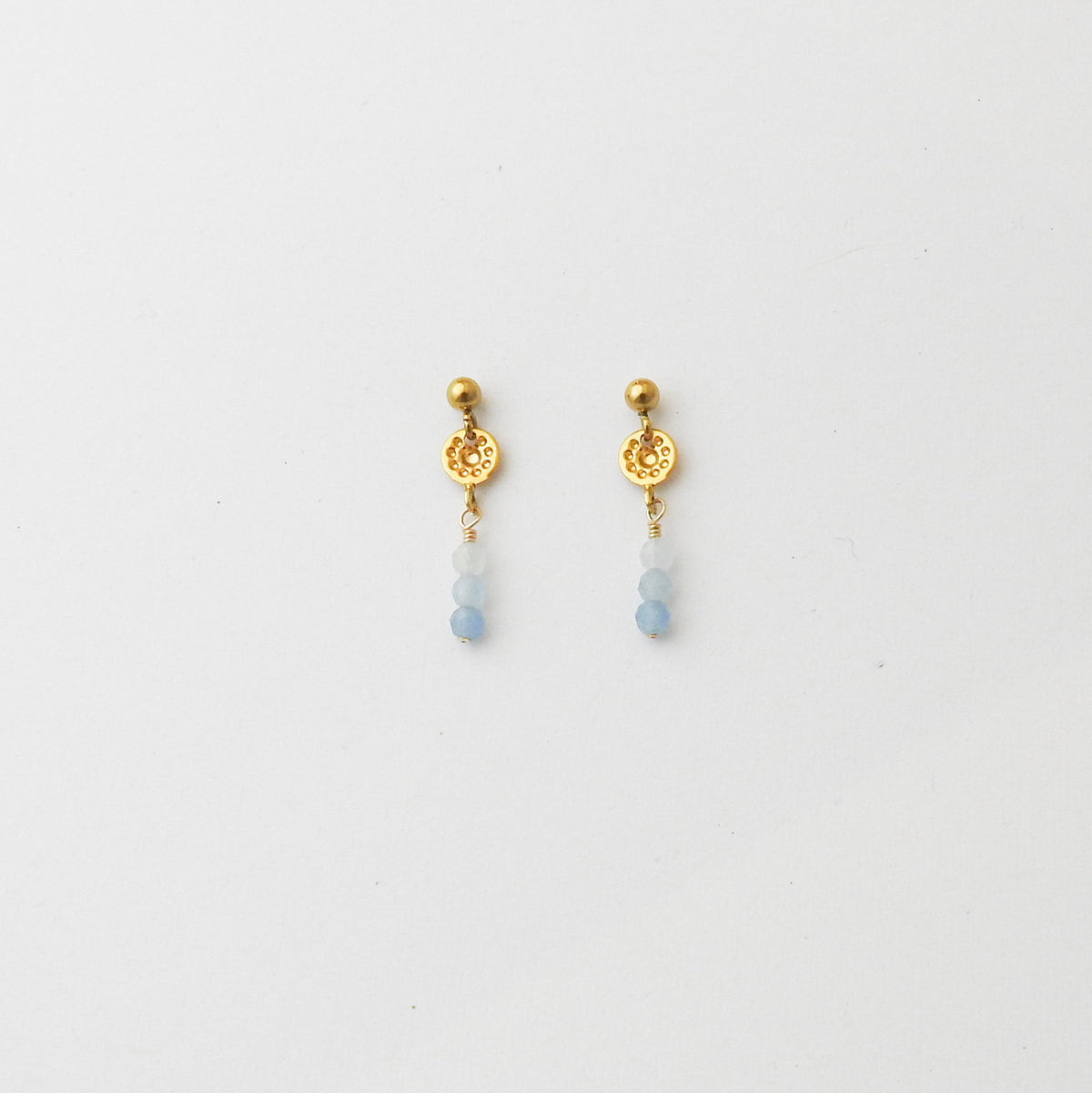 A pair of gold dangle earrings with a textured circular disc, a small gold bead, and three light blue beads hanging below, shown on a plain white background.