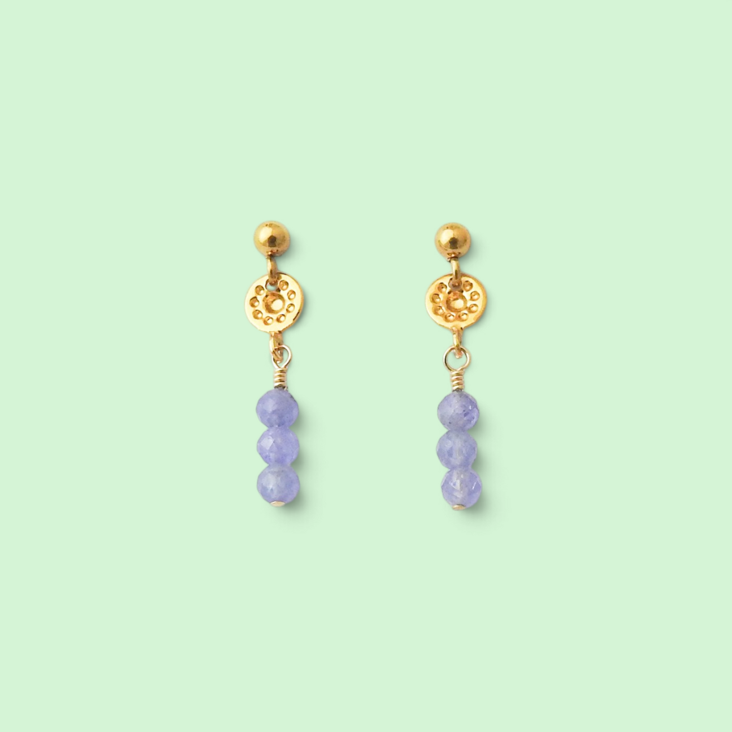 A pair of gold dangle earrings with a textured circular disc, a small gold bead, and three faceted light purple beads hanging below, set against a solid pale mint green background.