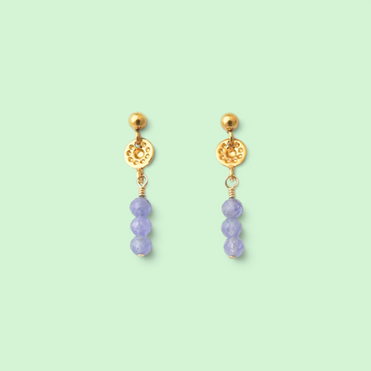 A pair of gold dangle earrings with a textured circular disc, a small gold bead, and three faceted light purple beads hanging below, set against a solid pale mint green background.