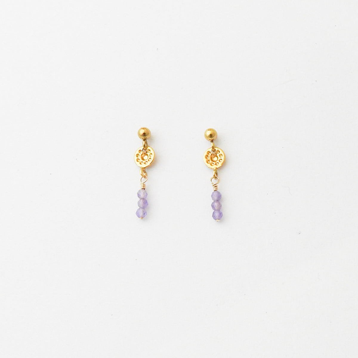 A pair of gold-toned dangle earrings with a textured circular charm and three small light purple beads hanging below, shown on a plain white background.