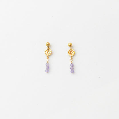 A pair of gold-toned dangle earrings with a textured circular charm and three small light purple beads hanging below, shown on a plain white background.