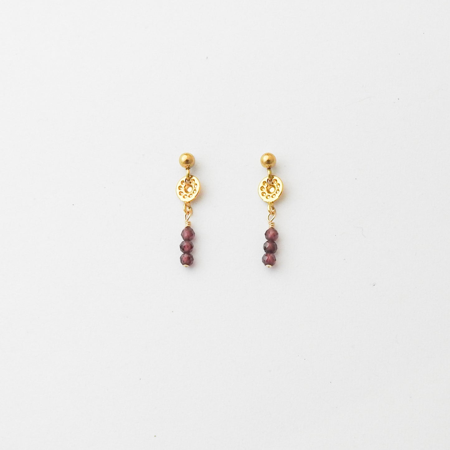 A pair of gold dangle earrings with a circular filigree disc, a small gold bead, and three dangling faceted dark red beads, shown on a plain white background.