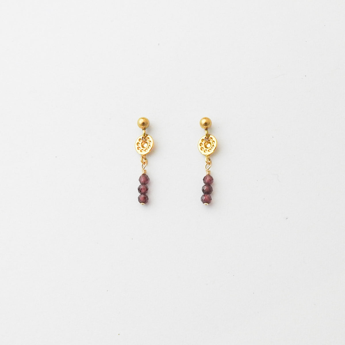 A pair of gold dangle earrings with a circular filigree disc, a small gold bead, and three dangling faceted dark red beads, shown on a plain white background.